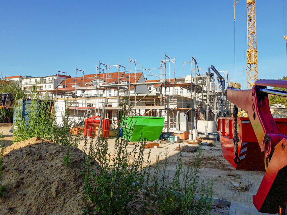 Die Baustelle von weiter weg. Davor ein aufgeschütteter Erdhügel, auf dem einige Pflanzen wuchern