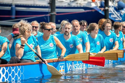 In einem großen Boot auf dem Wasser sitzen Menschen mit Paddeln und warten