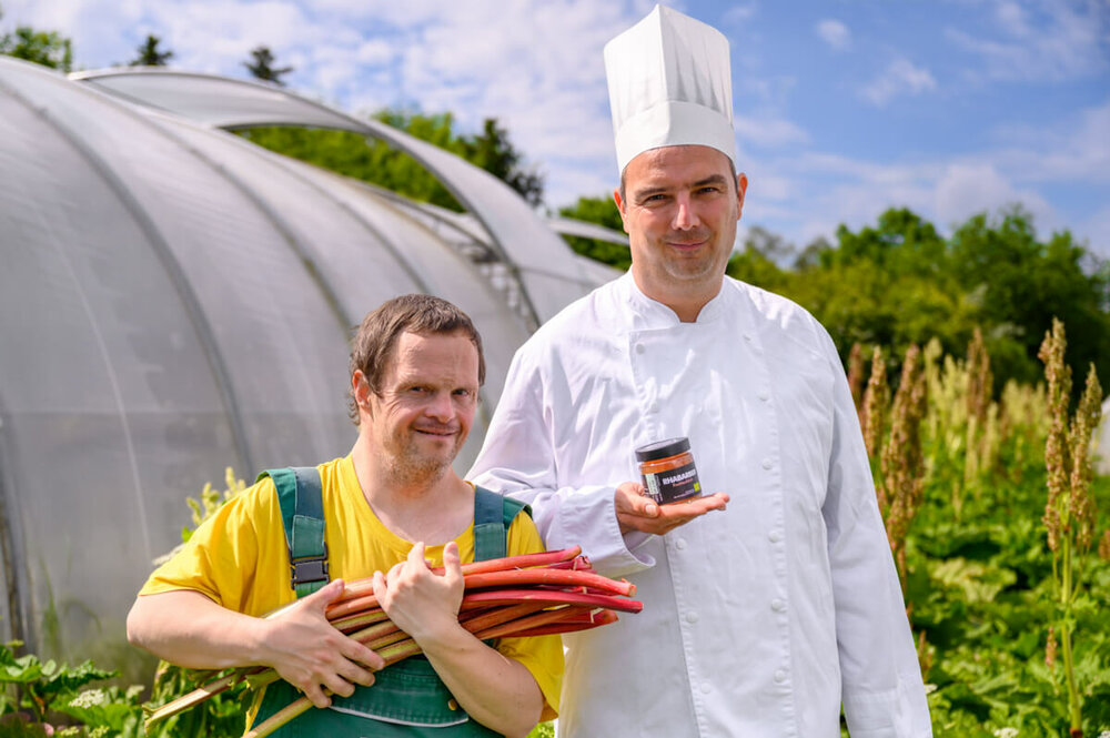 Zwei Männer stehen vor einem Gewächshaus und präsentieren frisches Gemüse und ein Glas mit Aufstrich
